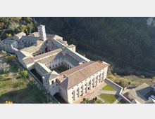 Veduta aerea di un grande complesso monastico in pietra con tetti in coppi e una torre campanaria, organizzato attorno a un chiostro interno. L’edificio sorge ai margini di un’area boscosa su un pendio, con cortili e spazi verdi perimetrali.