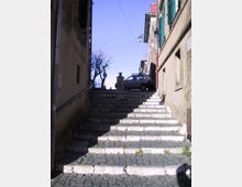 Scalinata in pietra e selciato che sale tra due edifici stretti, con ombre marcate sui gradini. In cima si vedono un lampione, un piccolo muretto in pietra e una strada con un’auto parcheggiata, sotto un cielo sereno.