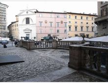 Veduta di una piazza urbana con pavimentazione in pietra e una terrazza rialzata con balaustra in muratura. Sullo sfondo si vedono una chiesa con facciata bianca e edifici storici color pastello, oltre a una strada con traffico e strutture coperte tipo dehors.
