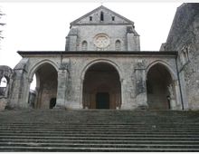 Facciata in pietra di una chiesa storica con ampio portico a tre arcate e rosone centrale, raggiungibile tramite una lunga scalinata. Ai lati si vedono muri in pietra e un arco d’accesso all’area esterna.