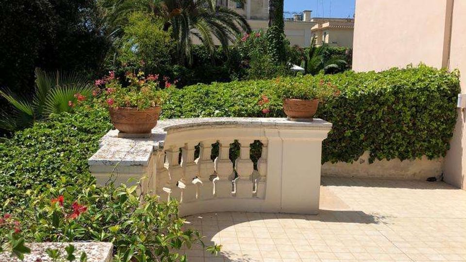 Terrazza piastrellata con balaustra in pietra chiara e vasi di fiori, affacciata su un giardino con siepi e alte palme. Sullo sfondo si vedono edifici chiari e cielo sereno.