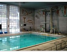 Piscina coperta con grande vasca rettangolare e piattaforme per tuffi su una torre, circondata da bordo in cemento e corrimano. Alte finestre a griglia illuminano l’ambiente e sulle pareti sono dipinti murales con figure di tuffatori e nuotatori.