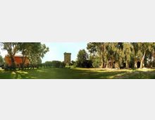 Ampio prato verde in un parco alberato, con una torre in pietra merlata al centro sullo sfondo. Ai lati si vedono un edificio in muratura rossastra e una struttura con archi, circondati da alberi alti.