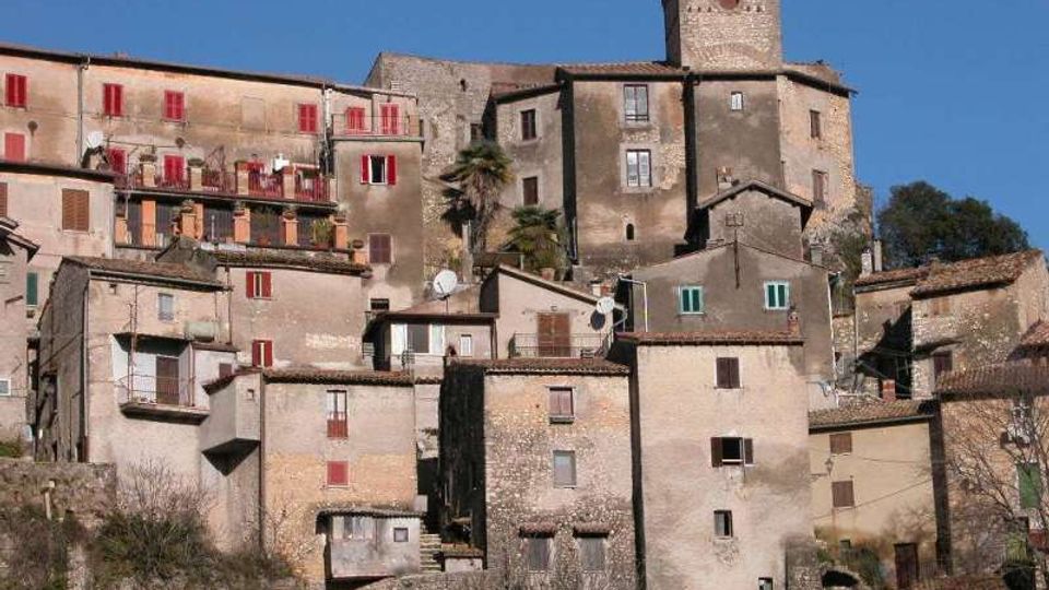 Borgo medievale su un pendio con case in pietra addossate tra loro, finestre e persiane colorate e tetti in coppi. In alto domina una torre quadrata merlata in pietra, con cielo sereno sullo sfondo.