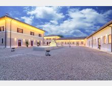 Cortile interno in ghiaia di un complesso edilizio con edifici bassi intonacati e tetti in tegole, illuminati da luci calde lungo le facciate. Al centro si trova una piccola fontana con muretto curvo, sotto un cielo parzialmente nuvoloso.