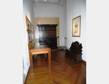 Interno di una piccola sala museale con pareti bianche e pavimento in parquet a spina di pesce, arredato con una grande credenza scura, una panca antica e una vetrina espositiva in legno e vetro. Sulle pareti sono appesi quadri incorniciati e l’illuminazione è a soffitto alto.