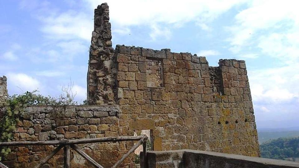 Rovine di una struttura in pietra con alti muri parzialmente crollati, affacciata su un paesaggio collinare. In primo piano si vede una terrazza con parapetto e una staccionata in legno, sotto un cielo parzialmente nuvoloso.