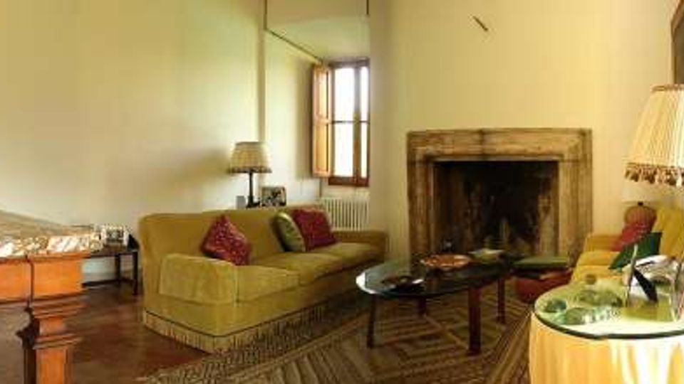 Salotto interno con divano verde, tavolino in vetro e grande camino in pietra, illuminato da lampade da tavolo e da una finestra. Arredi classici con credenza in legno, tappeto a motivi geometrici e porte in legno su pareti chiare.