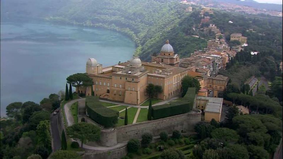 Veduta aerea di un grande complesso storico fortificato con cupole e giardini geometrici terrazzati, circondato da mura e alberi. Sullo sfondo si vedono un lago lungo la costa e un borgo collinare con edifici addossati al pendio.