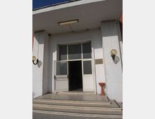 Ingresso di un edificio con facciata bianca e portico sporgente, raggiungibile tramite alcuni gradini. Porta a due ante con vetri opachi e sopraluce, lampade a parete ai lati e una piccola targa sulla destra.