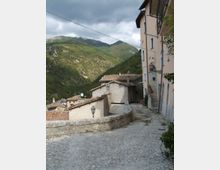 Vicolo acciottolato in un borgo di montagna, con case in pietra intonacata e tetti in coppi, muretto curvo e lampione. Sullo sfondo si vedono colline boscose e montagne sotto un cielo nuvoloso.