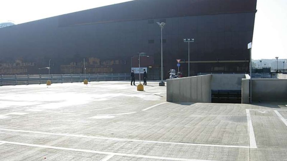 Ampia area pavimentata tipo parcheggio o piazzale, con segnaletica a linee bianche, lampioni e una rampa di accesso in cemento. Sullo sfondo si vede un grande edificio moderno scuro con facciata inclinata e superfici lisce.