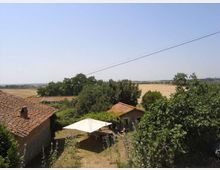 Vista di una casa di campagna con tetti in coppi e un cortile con gazebo bianco, circondata da alberi. Sullo sfondo si estendono campi coltivati e colline sotto un cielo limpido, con cavi elettrici che attraversano la parte alta dell’immagine.