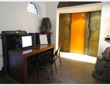Interno di una sala museale con pavimento in pietra, scrivania in legno con computer e sedie davanti a un mobile scuro. Sul fondo si vede un grande portale con pannello centrale arancione incassato in pareti metalliche, con illuminazione a soffitto e alcune sculture esposte ai lati.