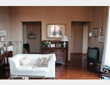Soggiorno domestico con pavimento in parquet e pareti color rosa tenue, arredato con un divano bianco, una cassettiera in legno con cornici e lampada, e un televisore su supporto nell’angolo. Due aperture interne conducono ad altre stanze e quadri incorniciati decorano le pareti.
