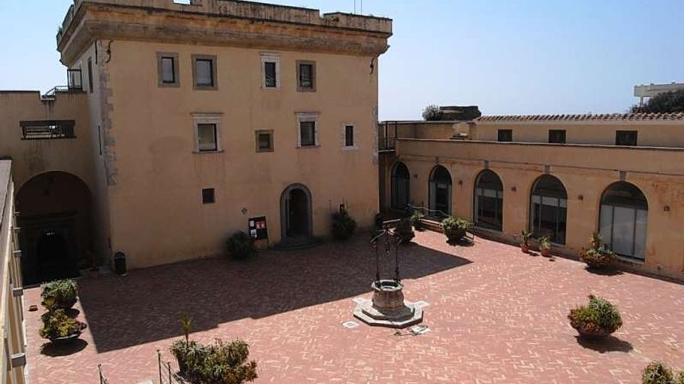 Cortile interno pavimentato in mattoni rossi, con un pozzo in pietra al centro e alcune fioriere. Sullo sfondo si vedono edifici color ocra con finestre rettangolari e un porticato ad archi lungo il lato destro.
