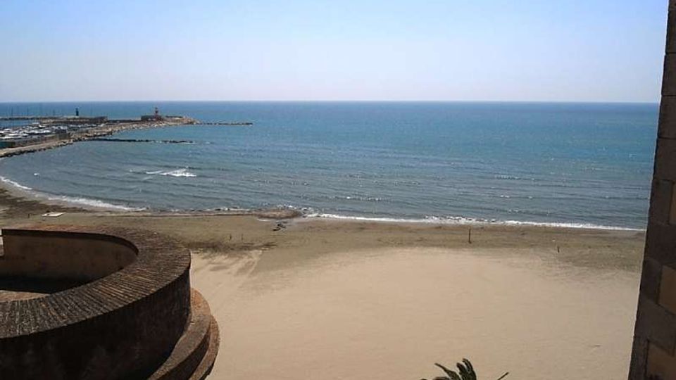Veduta dall’alto di una spiaggia sabbiosa e del mare calmo, con frangiflutti e un molo che si estendono verso l’orizzonte. In primo piano si vede una struttura in muratura curva e un parapetto in mattoni.