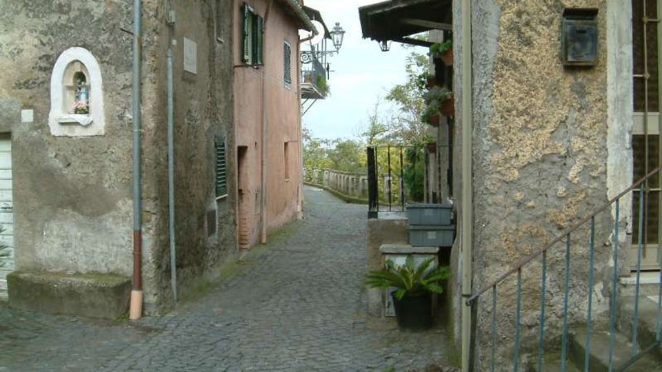 Vicolo stretto in pietra con pavimentazione in ciottoli, fiancheggiato da edifici in muratura intonacata e in pietra con persiane e piccoli balconi. In fondo la strada curva verso una ringhiera con vista su alberi, con lampioni e dettagli come una nicchia sul muro e vasi vicino a una scalinata.