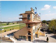 Edificio principale di un ippodromo, a più piani, con facciata in intonaco chiaro, balconi e terrazze, affacciato su una pista di corsa con prato e ringhiere. Intorno si vedono gradinate, scale esterne, strutture tecniche e alberi, con cielo sereno sullo sfondo.