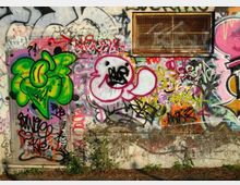 Parete esterna in cemento ricoperta da numerosi graffiti e scritte multicolori, con un disegno verde e una figura stilizzata rosa al centro. In alto a destra è presente una grata rettangolare su una finestra o presa d’aria, con vegetazione bassa alla base del muro.