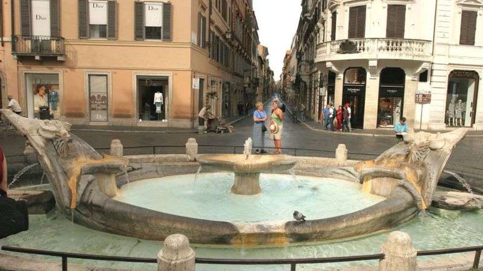 Fontana in pietra con vasca circolare e quattro sculture a forma di delfino che versano acqua, situata al centro di un incrocio tra strade lastricate. Sullo sfondo si vedono palazzi storici a più piani con persiane e negozi al piano terra lungo una via urbana stretta.