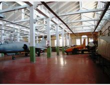 Ampio hangar industriale con pavimento rosso, colonne bianche e struttura del tetto a capriate con travi a vista. All’interno sono presenti diversi aerei e parti di velivoli, con grandi finestre sul fondo che illuminano lo spazio.