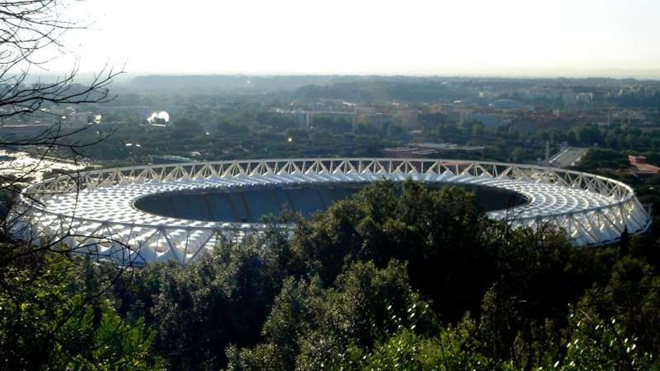 Veduta panoramica di un grande stadio ovale con copertura bianca a struttura reticolare, visto dall’alto tra alberi e vegetazione in primo piano. Sullo sfondo si estende la città con edifici e strade sotto un cielo luminoso.