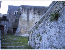 Cortile interno in pietra con gradini irregolari che salgono tra muri alti e scrostati, con tracce di umidità e vegetazione spontanea. Sullo sfondo si vede un edificio in muratura con piccole finestre ad arco e un passaggio scuro.