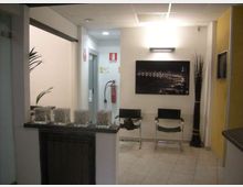 Piccola sala d’attesa interna con pavimento piastrellato e soffitto a pannelli, due sedie nere contro la parete e un quadro fotografico sopra. A sinistra si vede un bancone/reception con contenitori in vetro, mentre sul fondo ci sono una porta, un estintore a parete e un vaso alto con rami decorativi.