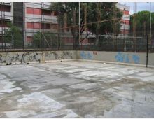 Cortile o area pavimentata in cemento con pozzanghere, circondata da muri bassi con graffiti e una recinzione metallica. Sullo sfondo si vedono alberi e un edificio residenziale a più piani con balconi e tende rosse.