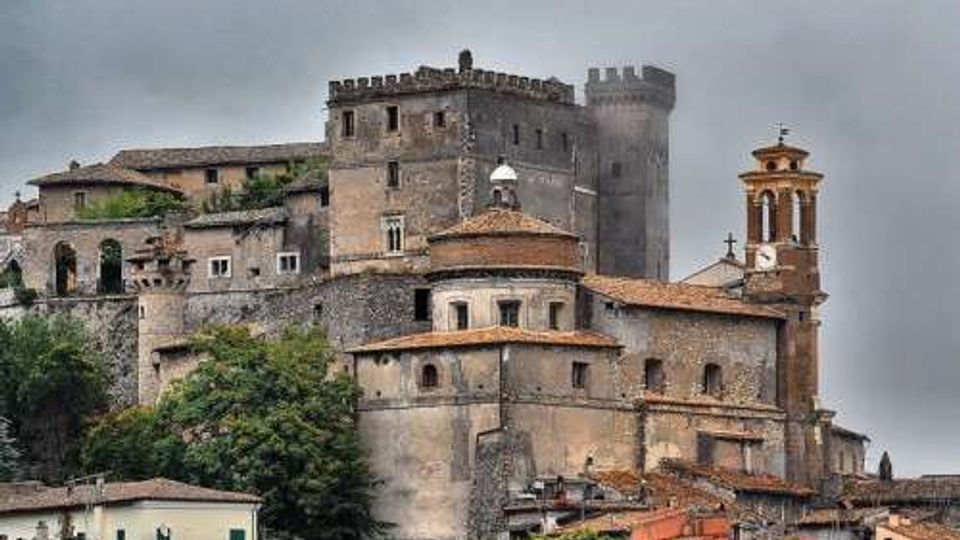 Castello medievale in pietra su un’altura, con mura merlate, una torre cilindrica e vari edifici annessi dai tetti in tegole. In primo piano si vedono case del borgo e, sul lato destro, un campanile con cuspide.