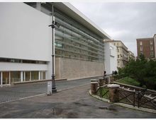 Edificio moderno con ampia facciata vetrata a pannelli orizzontali e volumi bianchi, affacciato su una strada pavimentata con lampione. In primo piano si vede un vialetto curvo con parapetto in pietra e legno che costeggia un’area verde, con palazzi urbani sullo sfondo.