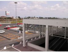 Area esterna di un grande centro commerciale con ampio parcheggio e corsie di accesso segnalate, con auto e lampioni alti. In primo piano una struttura moderna bianca con pilastri e pannelli orizzontali copre una rampa che scende verso un livello sotterraneo.
