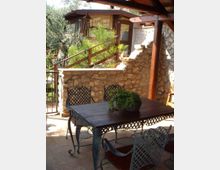Terrazza coperta con travi in legno, arredata con tavolo e sedie in ferro battuto su pavimento in cotto. Sullo sfondo si vede un muretto in pietra con vegetazione e una costruzione con pareti in pietra e grandi finestre affacciate sul giardino.