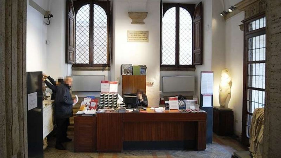 Interno di una biglietteria o reception in un edificio storico, con grande banco in legno al centro e materiali informativi esposti. Sullo sfondo ci sono due alte finestre ad arco con inferriate, pareti bianche con illuminazione a binario e una statua in una nicchia accanto a una porta vetrata.