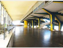 Ampio atrio interno di una struttura sportiva, con grande pavimentazione scura e colonne e travi in acciaio verniciate blu e giallo. Sulla sinistra si estende una parete di vetrate continue, mentre a destra si vedono gradinate e scale metalliche che conducono ai livelli superiori.