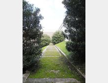 Vialetto in pietra e muschio che attraversa un’area verde in lieve pendenza, fiancheggiato da siepi e alberi. Sullo sfondo si intravedono edifici e un muro chiaro ai lati del percorso.