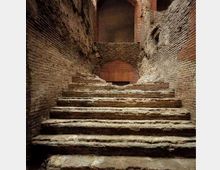 Scalinata in pietra consumata che sale tra strette pareti di mattoni e muratura antica. In cima si vede un arco in laterizio e un passaggio parzialmente chiuso da una muratura.
