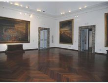 Sala museale con pareti bianche e pavimento in parquet scuro a spina di pesce, illuminata da faretti a soffitto. Grandi dipinti in cornici dorate sono appesi alle pareti e si vedono porte azzurre incorniciate in pietra che conducono ad altre stanze.
