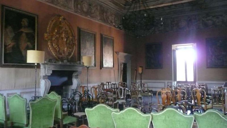 Sala storica con soffitto a cassettoni decorato e grande lampadario centrale, pareti con boiserie, dipinti e un camino in pietra. Numerose sedie d’epoca (anche con rivestimento verde) sono disposte in file, con una finestra luminosa sul fondo.