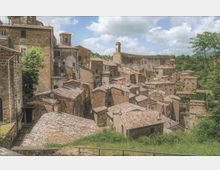 Vista panoramica di un borgo medievale su un pendio, con case in pietra addossate tra loro e tetti in coppi. Sullo sfondo si vedono una chiesa con campanile e una lunga muraglia sopra un’area verde collinare.