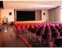 Sala auditorium con file di sedie imbottite rosse e nere disposte verso un palco rialzato con sipario nero. Soffitto con faretti incassati, pavimento lucido e grandi finestre laterali con tende che illuminano l’ambiente.