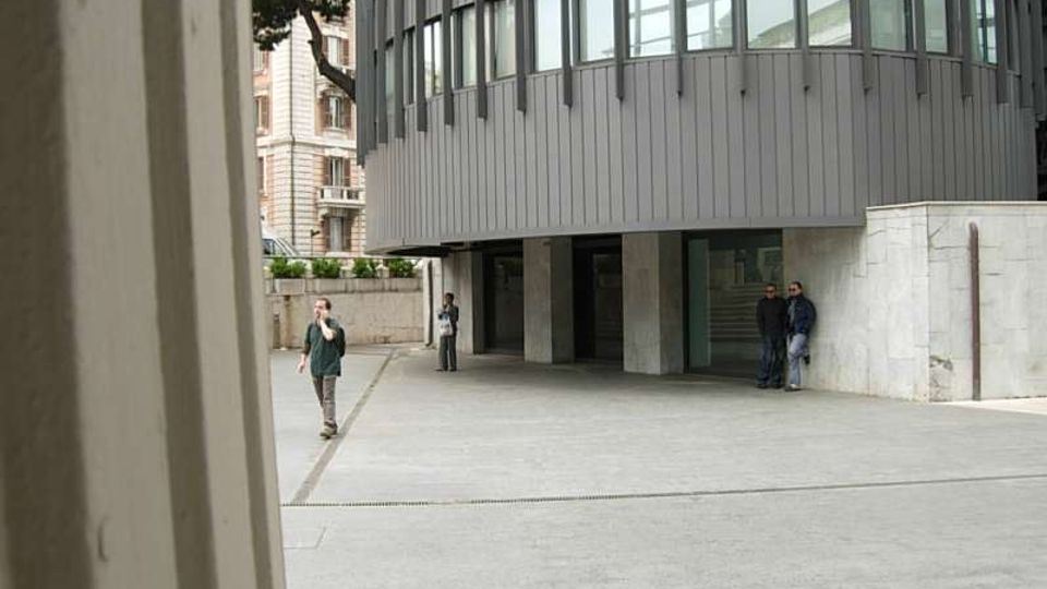 Piazzale urbano pavimentato davanti a un edificio moderno con facciata curva in pannelli metallici e ampie finestre, rialzato su pilastri con ingresso vetrato. Sullo sfondo si vedono un muro in pietra chiara, alberi e palazzi storici adiacenti.