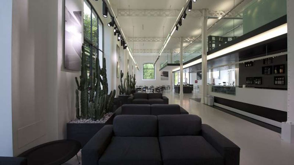 Interno di una sala lounge moderna con divani scuri allineati, grandi finestre laterali e fioriere con cactus lungo la parete. A destra si vede un lungo bancone bar con scaffali, illuminazione a binario e soffitto alto con travi a vista.