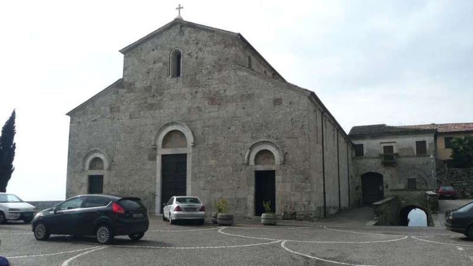 Facciata di una chiesa in pietra con ingresso ad arco e piccole finestre, affacciata su una piazza lastricata con linee curve e alcune auto parcheggiate. Sul lato destro si vedono edifici annessi e un passaggio ad arco in muratura.