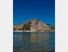 Veduta di una baia con acqua calma e riflessi in primo piano, con un promontorio roccioso e scogliere sullo sfondo. Alla base si vede un piccolo porto con barche a vela e un faro verde su un molo.