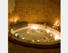 Vasca da bagno rotonda incassata in un bagno rivestito di piastrelle beige, con rubinetteria cromata e acqua. Candele accese e petali di rosa sono disposti sul bordo e sulla superficie dell’acqua, con asciugamani appesi sulla parete.