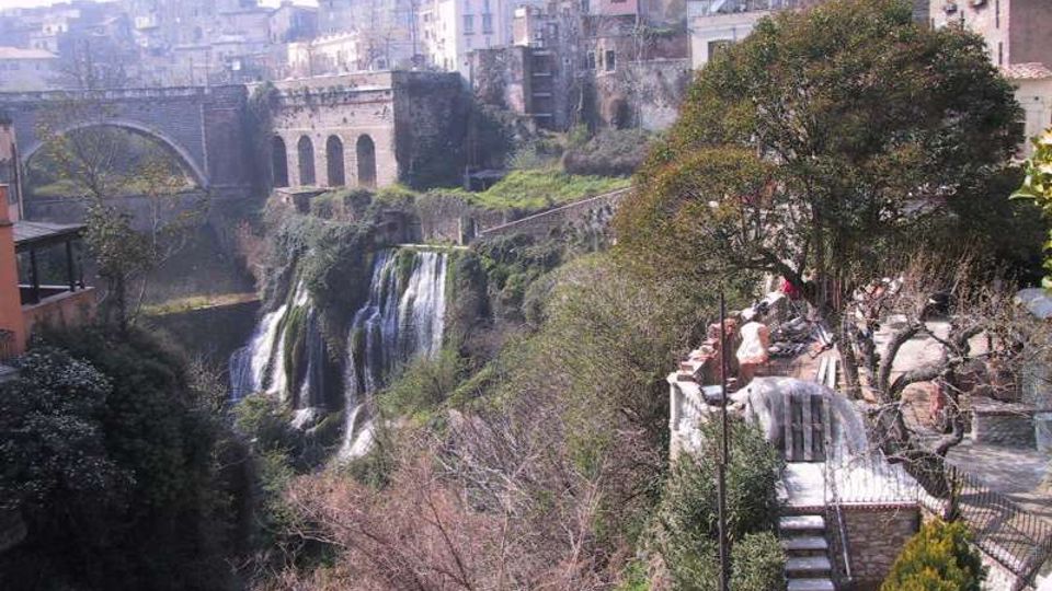 Veduta panoramica di un borgo storico su una gola, con edifici in pietra, un ponte ad arco e un antico viadotto sullo sfondo. In basso si vede una cascata che scende tra vegetazione fitta, con terrazze e scale lungo il versante sulla destra.