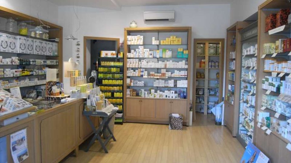 Interno di un negozio tipo farmacia/erboristeria con pavimento in legno chiaro, pareti bianche e scaffalature in legno lungo i lati e sul fondo piene di confezioni e prodotti. Al centro c’è un piccolo tavolo espositivo e sul fondo si vede una porta che conduce a un’area sul retro.