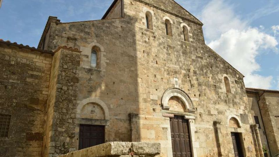 Facciata in pietra di una chiesa romanica con portale centrale ad arco e grandi porte in legno, affiancata da ingressi laterali. La struttura massiccia presenta piccole finestre ad arco in alto ed è ripresa dall’esterno sotto un cielo parzialmente nuvoloso.
